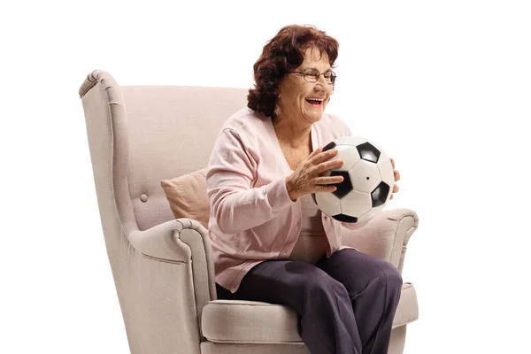 Senior woman sitting in an armchair with soccer ball and smiling — Zdjęcie stockowe