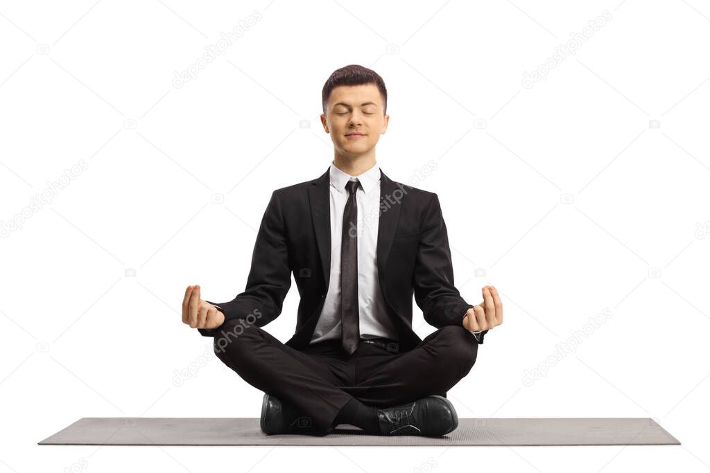 Young man in a black suit with closed eyes sitting with crossed legs and making a meditation pose on a mat isolated on white background