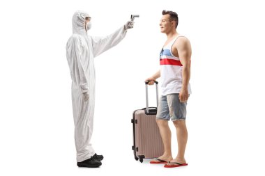 Tehlikeli madde giysisi giymiş bir adamın, beyaz arka planda izole edilmiş bir bavulu olan bir erkek turistin vücut ısısını ölçerken tam boy profil fotoğrafı.
