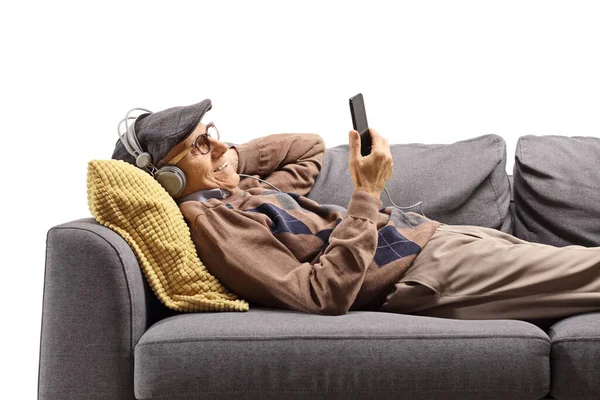 快乐的老年人躺在沙发上 手持手机 用白色背景的耳机听音乐 — 图库照片