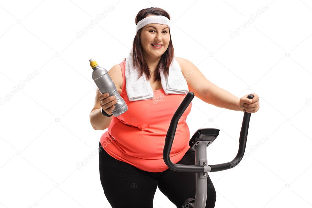 Chubby young woman on a stationary bike holding a plastic bottle isolated on white background