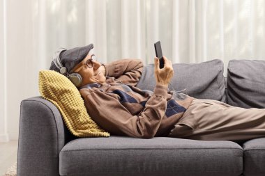 Kanepede oturan yaşlı bir adam cep telefonu tutuyor ve evde kulaklıkla müzik dinliyor.