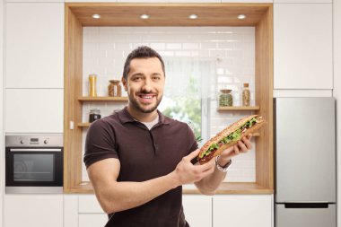 Genç adam mutfakta salatalı sandviç tutuyor. 