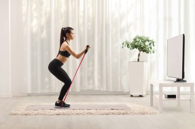 Spor giyimli genç bir kadın evde bir televizyon karşısında direniş bandıyla egzersiz yapıyor.