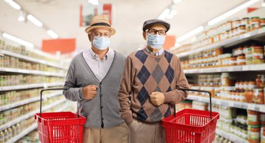 Tıbbi maske takan ve süpermarkette alışveriş sepetleri tutan iki kıdemli adam.