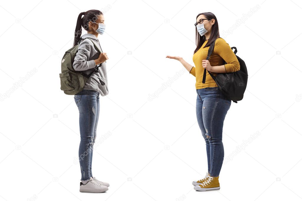Full length profile shot of a two female students with protective medical masks having a conversation isolated on white background