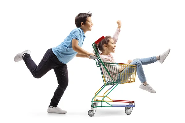 白い背景に隔離されたショッピングカートの中で女の子を押して小さな男の子の完全な長さのプロフィールショット — ストック写真