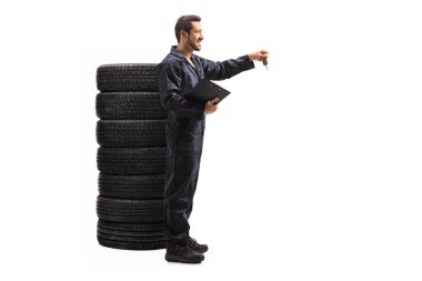 Anahtarları tutan bir otomobil tamircisinin tam uzunluktaki profil çekimi ve beyaz arka planda izole edilmiş bir lastik yığınının yanında durmak.