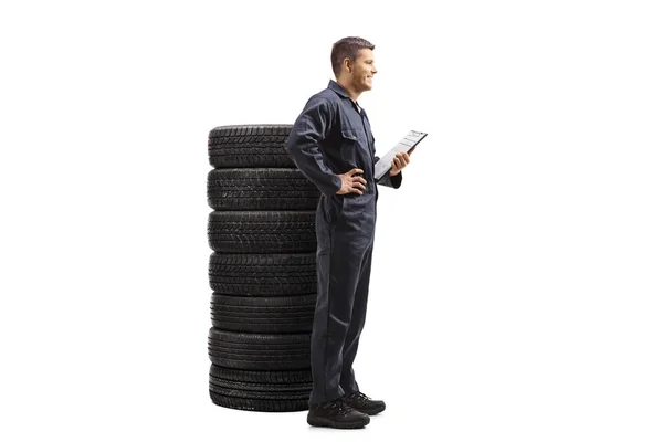 一个身穿制服的汽车修理工拿着剪贴板站在一堆轮胎旁边 与白色背景隔离的全长轮廓照片 — 图库照片
