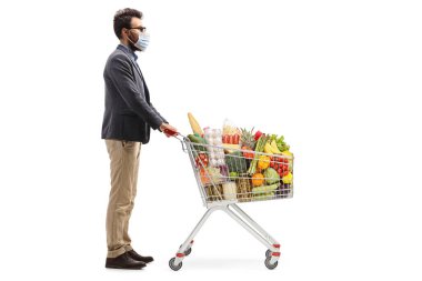 Sakallı bir adamın koruyucu yüz maskesi taktığı ve beyaz arka planda yiyecek dolu bir alışveriş arabasıyla dikilirken tam boy profil fotoğrafı.