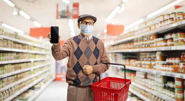 Senior man with a shopping basket in a supermarket, wearing a protective face mask and showing a mobile phone