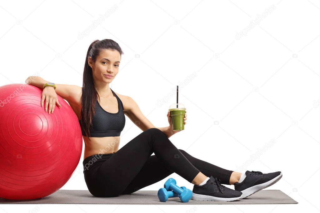Young female sitting on the floor with a fitness ball and a healthy green smoothie isolated on white background