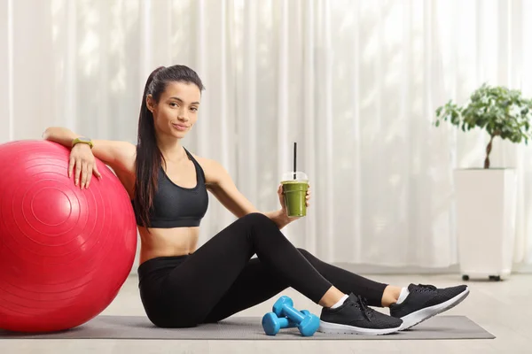 Fit woman sitting on the floor with a fitness ball and a healthy green smoothie at home