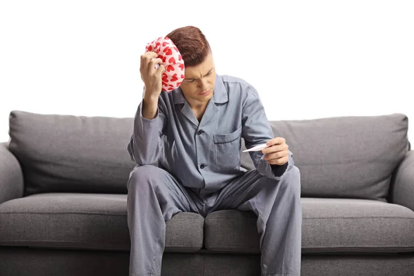 年轻人穿着睡衣坐在沙发上测量体温 头上拿着一个止疼垫 与白色背景隔离 — 图库照片