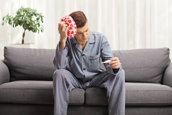 男座っているソファにパジャマで体温を測定し 彼の頭の上に痛み救済クッションを保持 — ストック写真