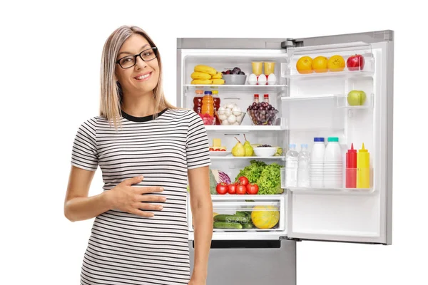 Молода Жінка Тримає Руку Животі Перед Повноцінним Холодильником Ізольовано Білому — стокове фото