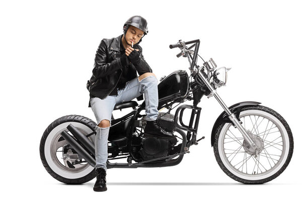 Young biker with a helmet and a leather jacket sitting on a chopper motorbike and lighting a cigarette isolated on white background