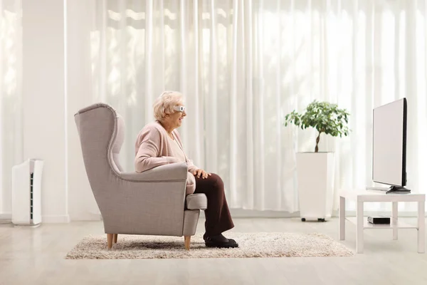 Elderly lady with a pair of 3D glasses sitting in an armchair and watching tv at home