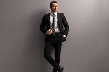 Gri bir duvara yaslanmış siyah takım elbiseli bir erkek model. 