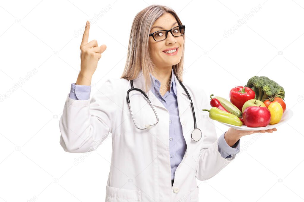 Female doctor holding a plate of fresh fruits and vegetables and pointing up isolated on white background