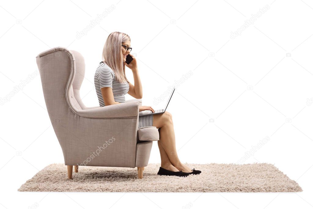 Woman sitting in an armchair and working from home on a laptop computer while talking on a phone isolated on white background