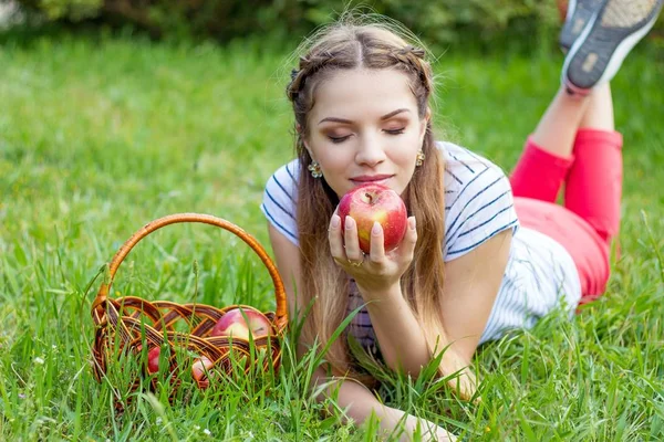 Молода красива жінка лежить на зеленій траві. Дівчина і кошик з червоних яблук. Тепла весна . — стокове фото