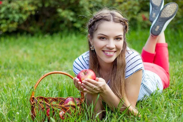 Jovem mulher bonita está deitada na grama verde. Uma menina e um cesto de maçãs vermelhas. Primavera quente . — Fotografia de Stock