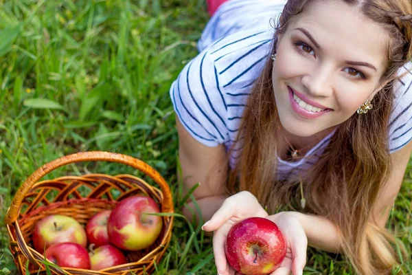 Jonge mooie vrouw ligt op het groene gras. Een meisje en een mandje van rode appels. Warm voorjaar. — Stockfoto