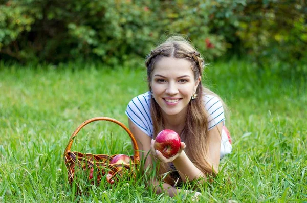 Молода красива жінка лежить на зеленій траві. Дівчина і кошик з червоних яблук. Тепла весна . — стокове фото