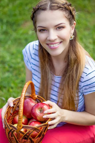 Jonge mooie vrouw ligt op het groene gras. Een meisje en een mandje van rode appels. Warm voorjaar. — Stockfoto