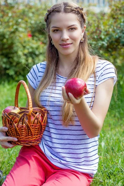 Jonge mooie vrouw ligt op het groene gras. Een meisje en een mandje van rode appels. Warm voorjaar. — Stockfoto