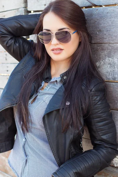 Mooie jongedame in zonnebril. Portret van een jonge vrouw op een houten achtergrond. Mooie brunette in zwarte jas. — Stockfoto