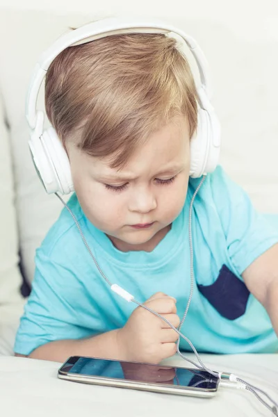 3-летний мальчик сидит на диване и слушает музыку. Маленький милый мальчик смотрит мультики по телефону. Маленький мальчик . — стоковое фото