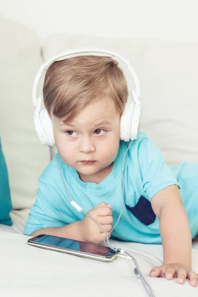 3-летний мальчик сидит на диване и слушает музыку. Маленький милый мальчик смотрит мультики по телефону. Маленький мальчик . — стоковое фото