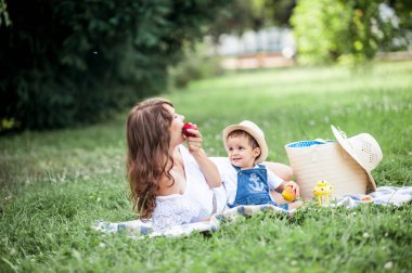 Anne ve genç oğlu çimenlerin üzerinde oynadı. Mutlu aile parkta yürüyor. Meyve yeme anne ve genç oğlu. Piknik. Yaz.