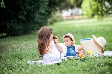 Anne ve genç oğlu çimenlerin üzerinde oynadı. Mutlu aile parkta yürüyor. Meyve yeme anne ve genç oğlu. Piknik. Yaz.