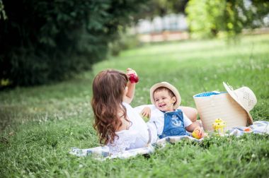Anne ve genç oğlu çimenlerin üzerinde oynadı. Mutlu aile parkta yürüyor. Meyve yeme anne ve genç oğlu. Piknik. Yaz.