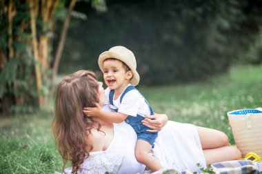 Anne ve genç oğlu çimenlerin üzerinde oynadı. Mutlu aile parkta yürüyor. Meyve yeme anne ve genç oğlu. Piknik. Yaz.