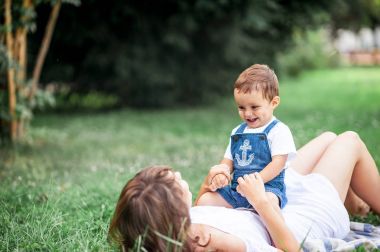 Anne ve genç oğlu çimenlerin üzerinde oynadı. Mutlu aile parkta yürüyor. Meyve yeme anne ve genç oğlu. Piknik. Yaz.