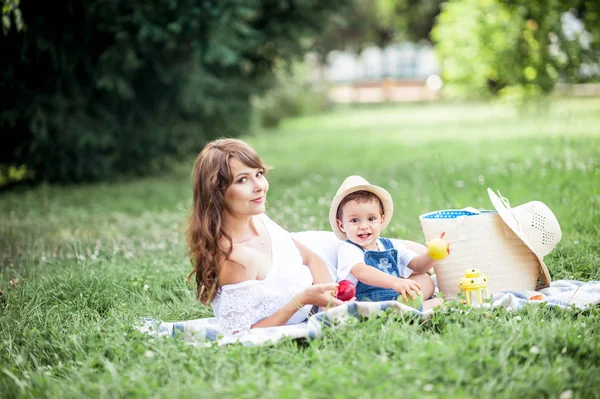 Matka i młody syn grał na trawie. Szczęśliwa rodzina spacery w parku. Mama i młody syn jedzenie owoców. Piknik. Latem. — Zdjęcie stockowe