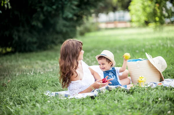 Matka i młody syn grał na trawie. Szczęśliwa rodzina spacery w parku. Mama i młody syn jedzenie owoców. Piknik. Latem. — Zdjęcie stockowe