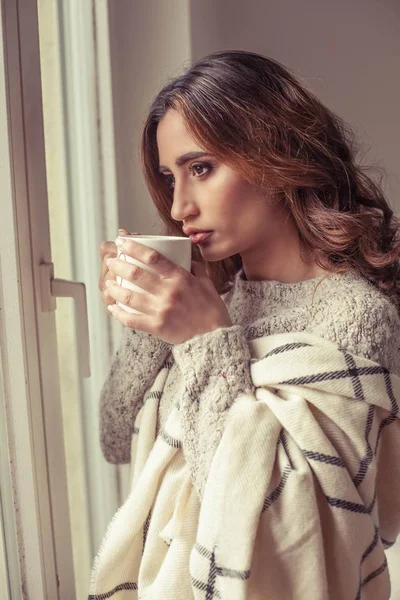 Young beautiful woman stands near a window, wrapped in a blanket and drink hot coffee. Cozy. Autumn. Warm cozy evening.