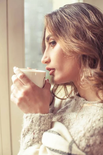 Joven hermosa mujer está cerca de una ventana, envuelta en una manta y beber café caliente. Acogedor. Otoño. Noche cálida y acogedora . — Foto de Stock
