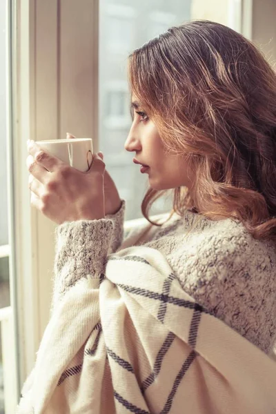 Unge, vakre kvinner står ved vinduet, svøpt i et teppe og drikker varm kaffe. Koselig. Høsten. Varm og koselig kveld . – stockfoto