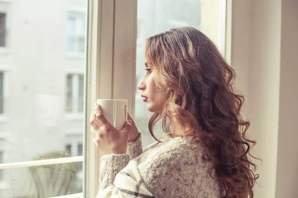 Unge, vakre kvinner står ved vinduet, svøpt i et teppe og drikker varm kaffe. Koselig. Høsten. Varm og koselig kveld . – stockfoto