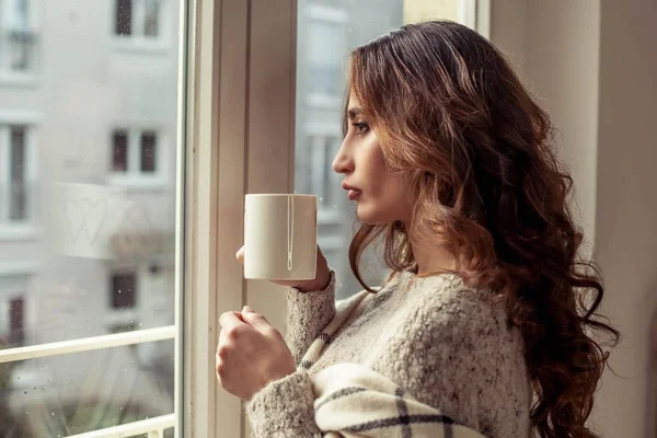 Unge, vakre kvinner står ved vinduet, svøpt i et teppe og drikker varm kaffe. Koselig. Høsten. Varm og koselig kveld . – stockfoto