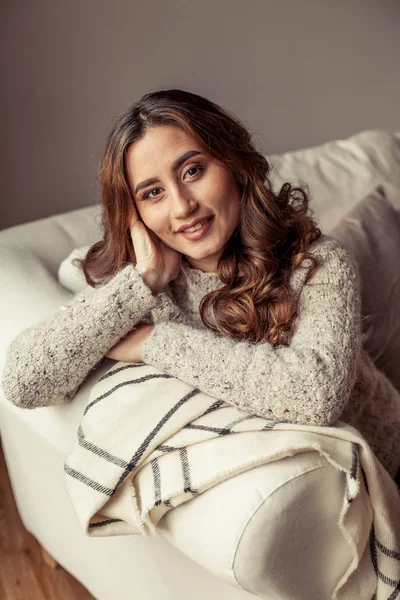 A cozy portrait of a young beautiful woman. Beautiful Turkish girl. Warm autumn. Cozy home evening. A young woman wrapped herself in a blanket and sits on the couch.