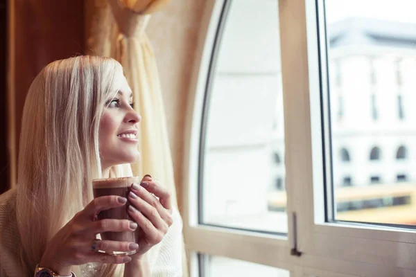 Las manos femeninas sostienen una taza de cacao caliente. Una joven está sentada en un acogedor café. La chica está bebiendo cacao. El invierno. Acogedor otoño . — Foto de Stock