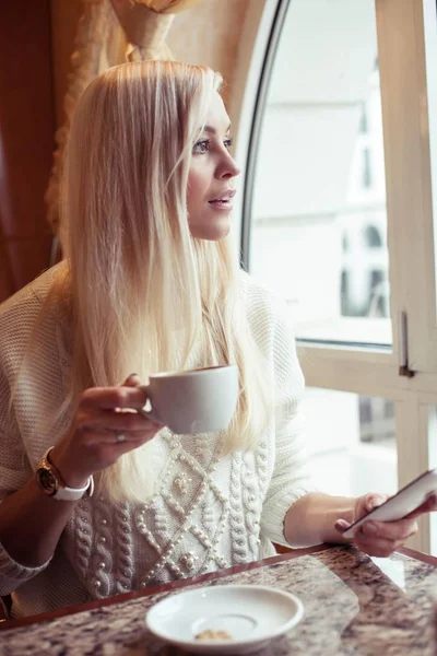 Una joven y hermosa mujer sostiene una taza de capuchino caliente. Una joven está sentada en un acogedor café bebiendo capuchino y hablando por teléfono. La chica está bebiendo cacao. El invierno. Acogedor otoño . — Foto de Stock