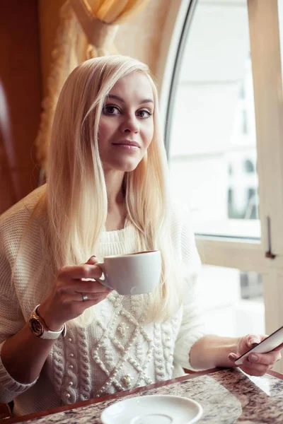 Una giovane bella donna sta tenendo una tazza di cappuccino caldo. Una giovane donna è seduta in un accogliente caffè a bere cappuccino e parlare al telefono. La ragazza sta bevendo cioccolata. Inverno. Autunno accogliente . — Foto Stock
